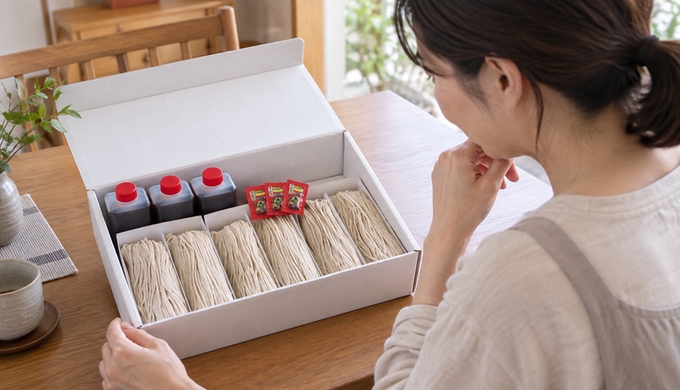 自宅のテーブルで、六束の生そばとつゆ、薬味が入った箱を開き、中身を見ている。