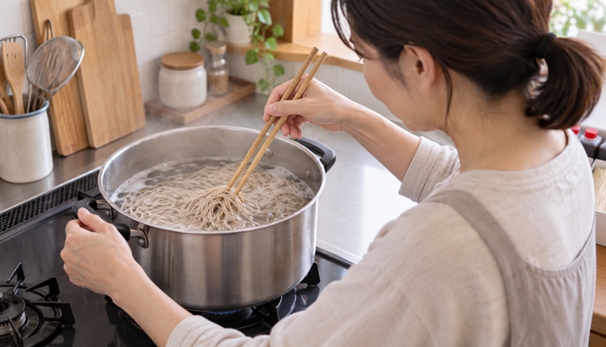 自宅のキッチンで、大きな鍋に入れた複数人分のそばを箸でゆでている。