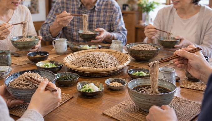 複数人分のそばが並ぶ食卓で、何人かが箸を伸ばして食べ始めている。
