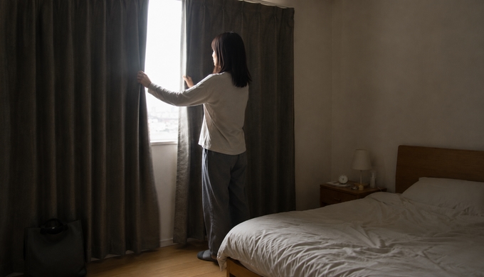 寝室の窓辺で女性が厚手のカーテンを閉め、朝の光が入る範囲を小さくしている。