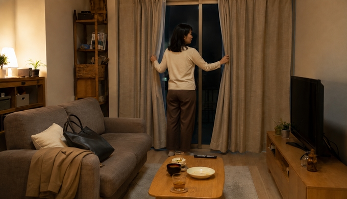 帰宅した女性が夜のリビングの窓辺でカーテンを閉めている。