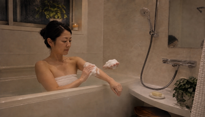 自宅の浴室で、女性が細かな石けんの泡を片手から前腕へゆっくり広げている。