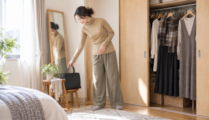 タートルネックとパンツを着た女性が、外出前に鏡のそばでバッグへ手を伸ばしている。