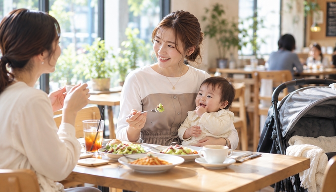 ビスチェ風ワンピースを着た女性が赤ちゃんと一緒にカフェの席につき、友人とメニューや料理を見ながら話している。